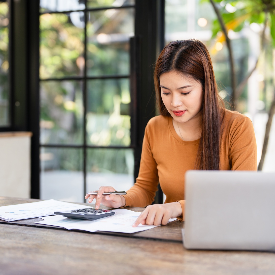 A woman sits at a table, reviewing documents while entering numbers into a calculator. Her laptop sits nearby.