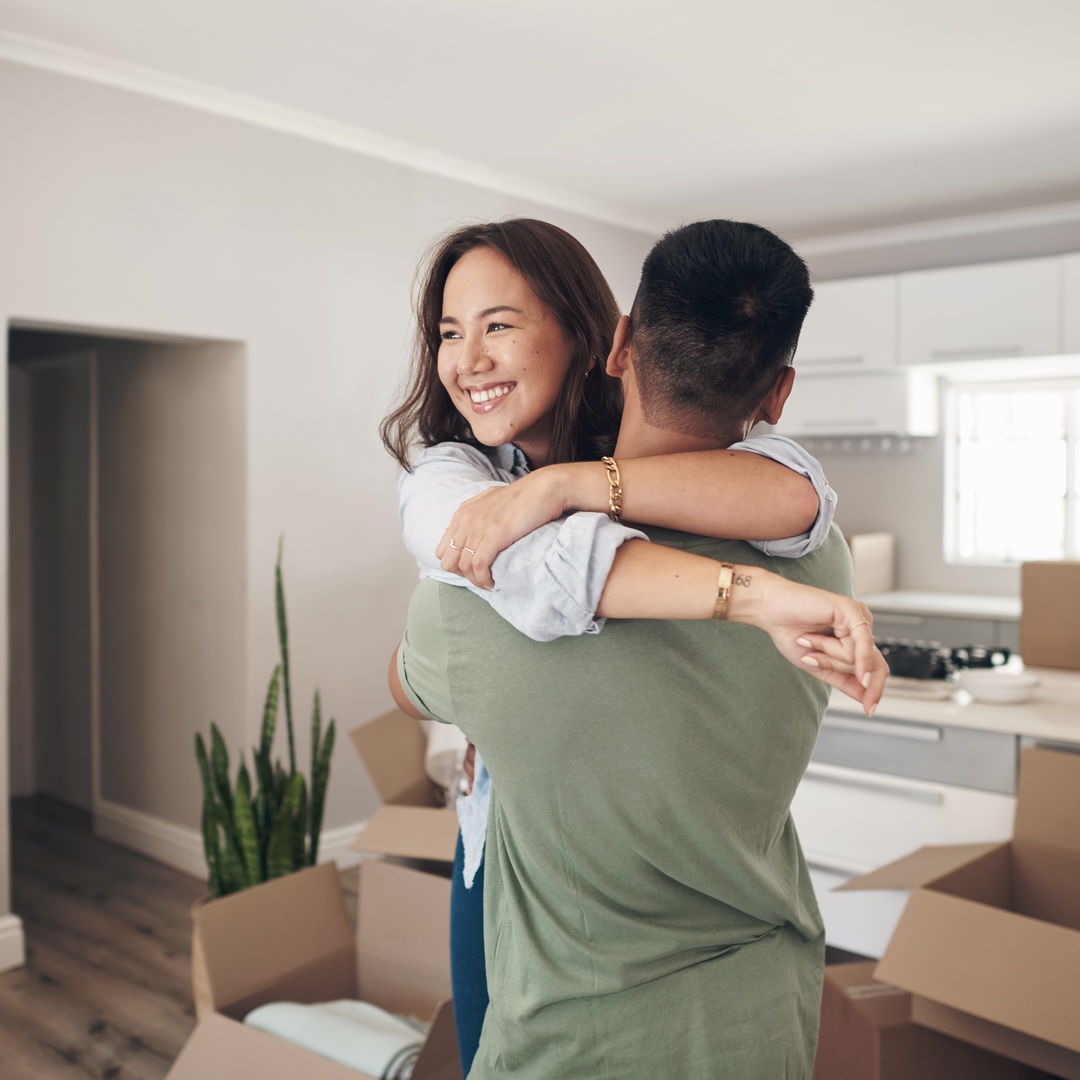 A smiling woman and a man hug in a spacious but empty kitchen full of cardboard boxes and white furniture.