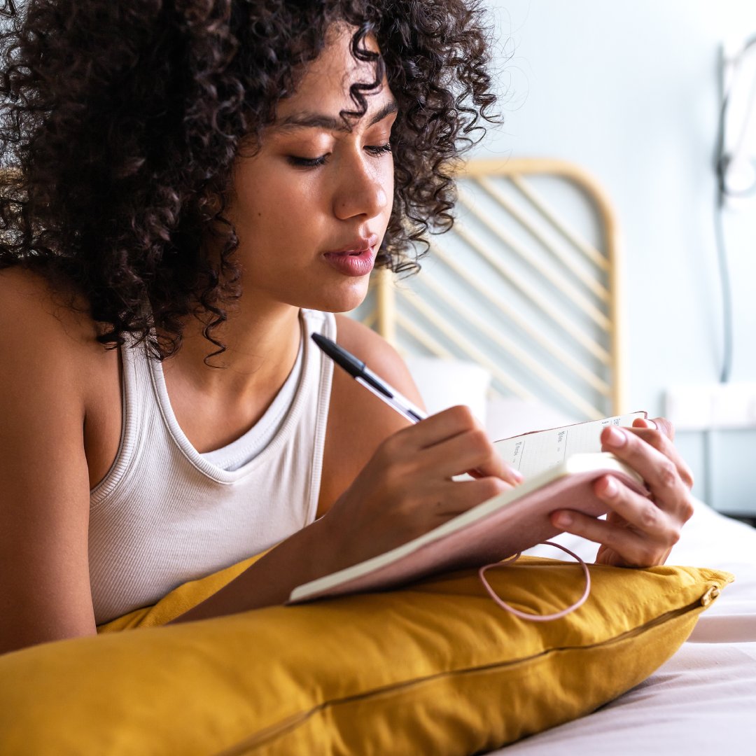 A woman lays on her stomach on top of her bed. She holds open a pink journal and writes something down.