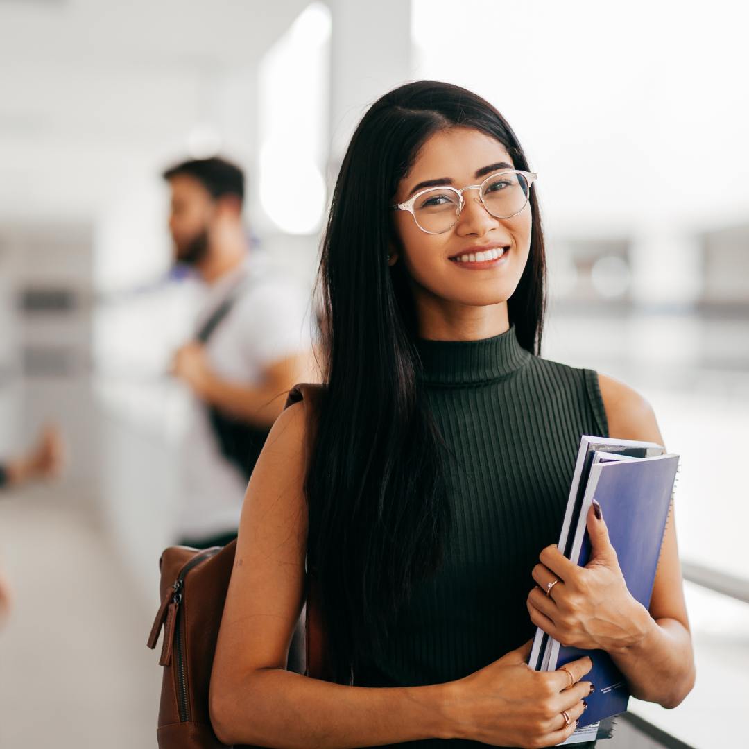 A young, studious woman is smiling as she walks to a college course. She's wearing a backpack and carrying a notebook.