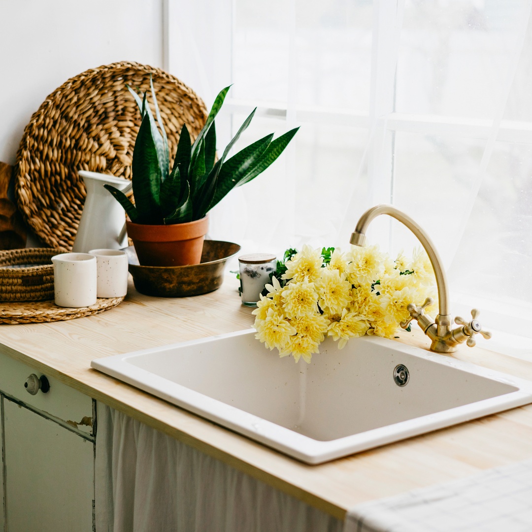 A counter with a farmhouse sink and a gold faucet. The corner has various boho inspired baskets, place mats, and a plant.