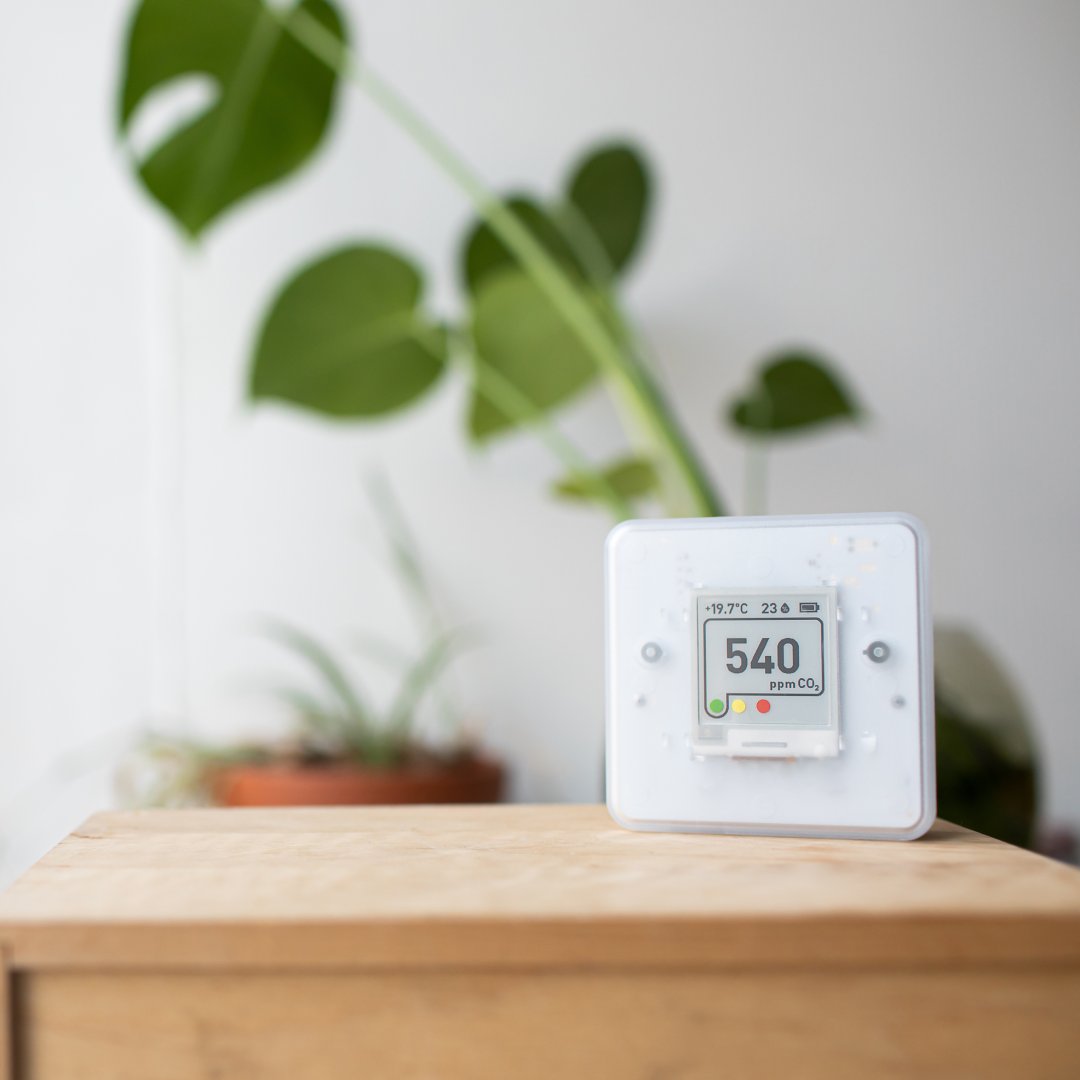 A white indoor air quality sensor sits on top of a wooden table. Behind the table are multiple potted plants.