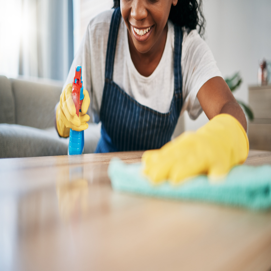 A woman wearing a blue apron and yellow gloves sprays a cleaning product on the table and wipes it down with a rug.