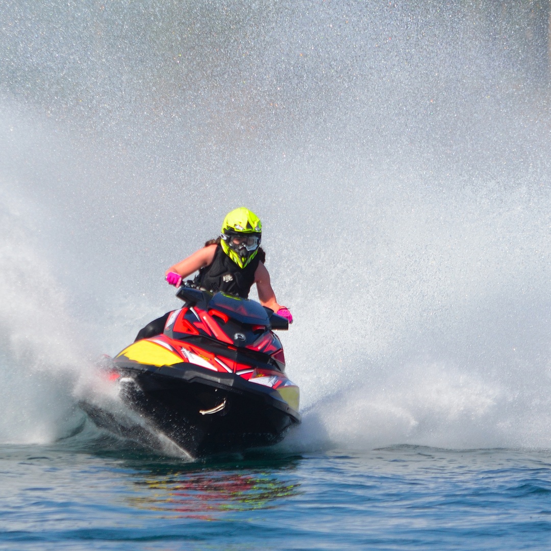 A person rides their jet ski on the water with a safety vest and a yellow helmet on. The water kicks up behind them.