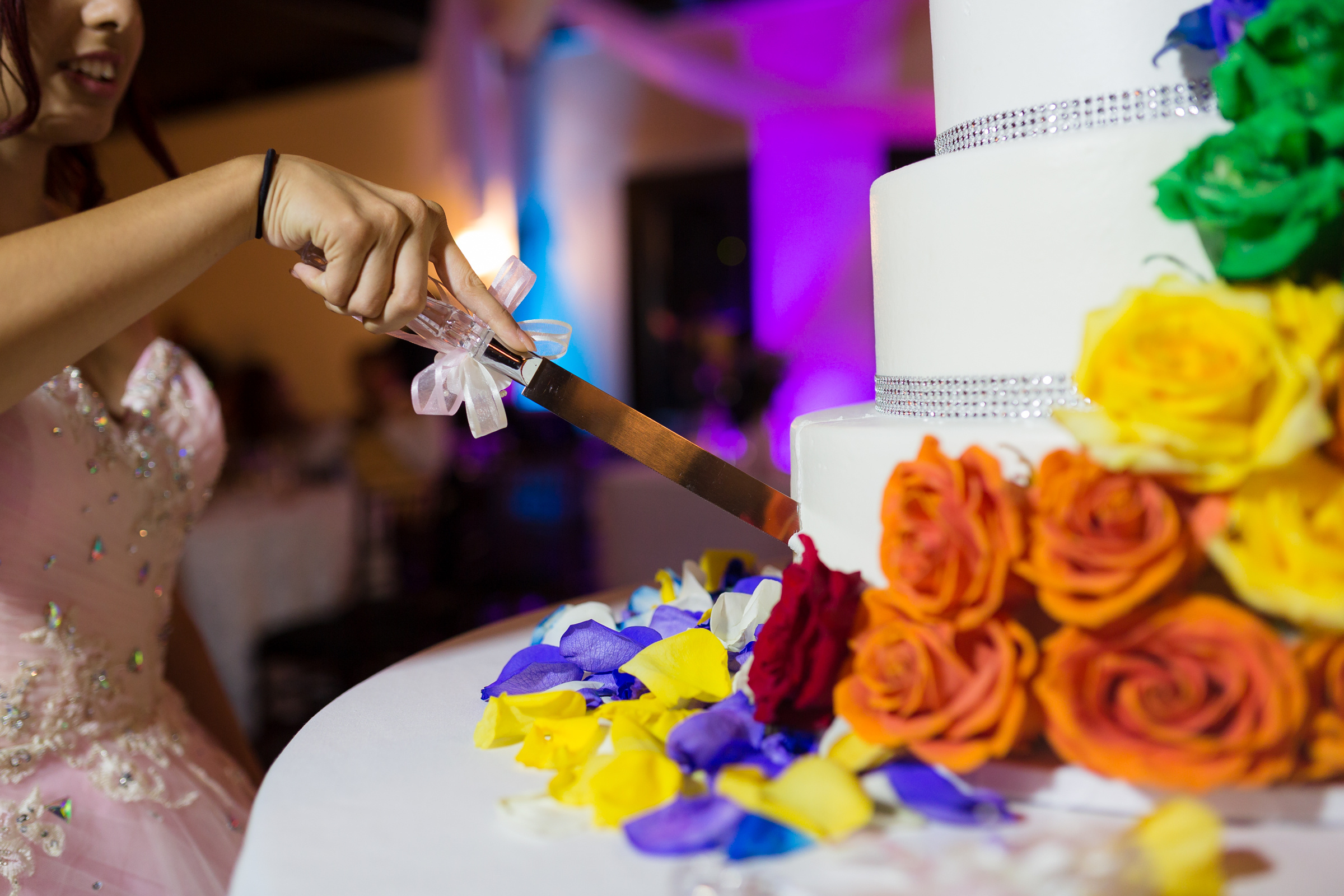 A girl in a pink bedazzled ball gown uses a long knife to cut a multi-tier cake with colorful flowers.