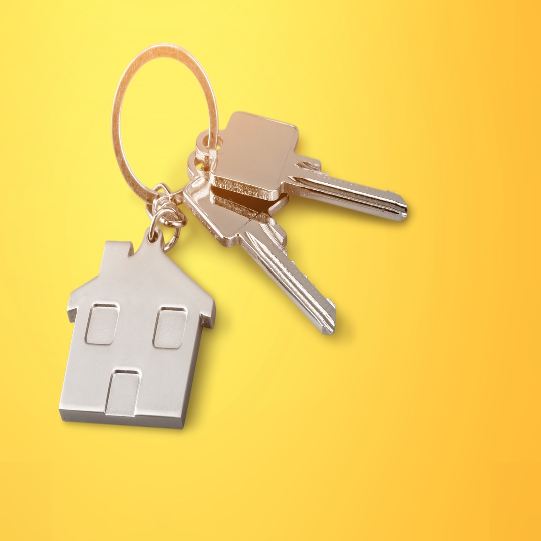 A metal keychain shaped like a house features windows and a door. It holds two keys and sits on a bright yellow background.