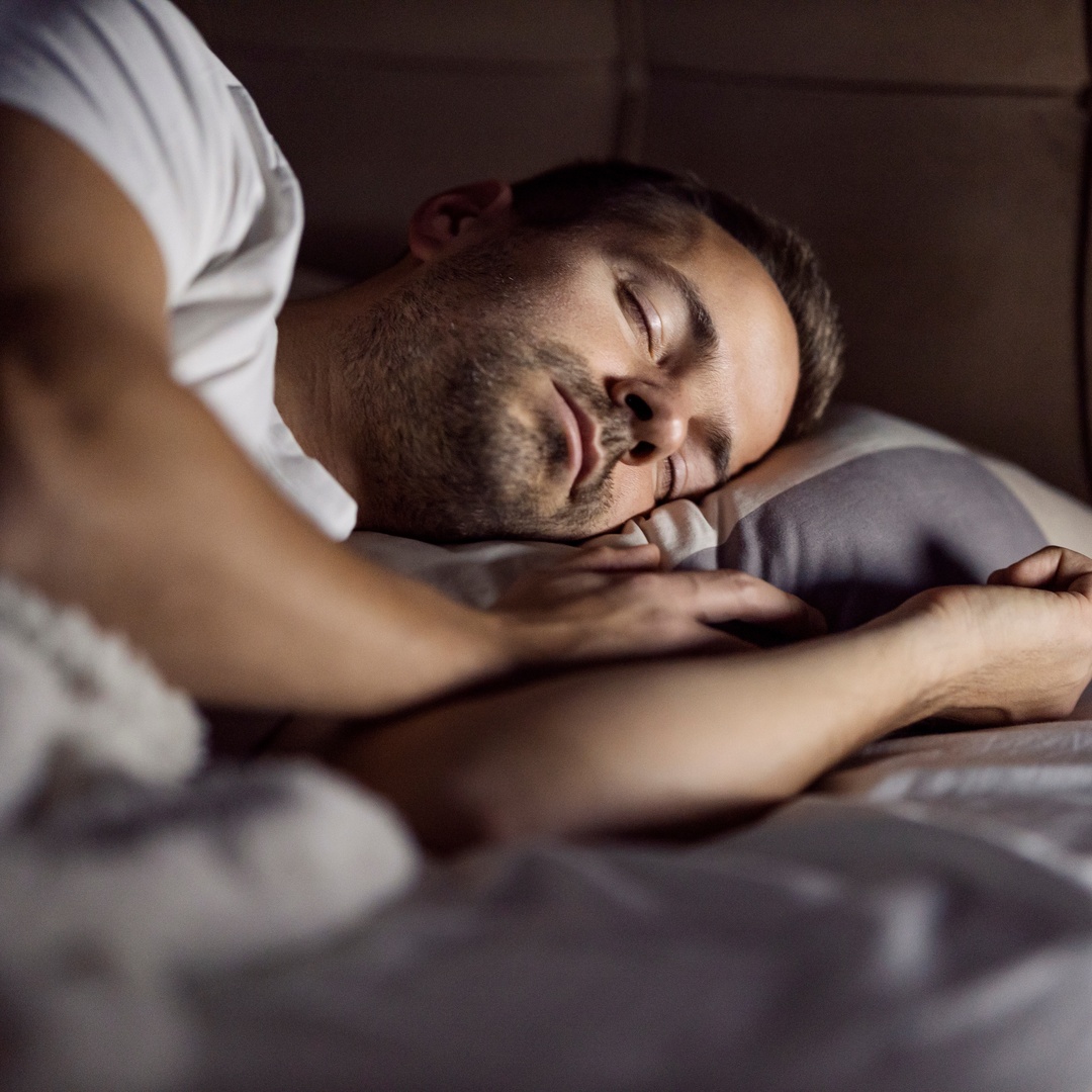 A man enjoys a deep sleep as he lies in bed. He wears a white t-shirt and rests his head on a cushy pillow.
