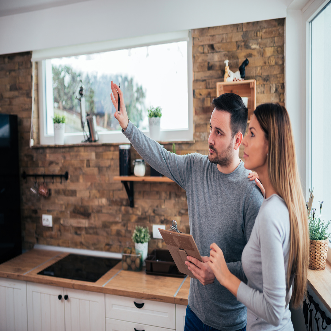 A man and a woman stands side by side while the man holds a clipboard and raise his hand up next to the kitchen.