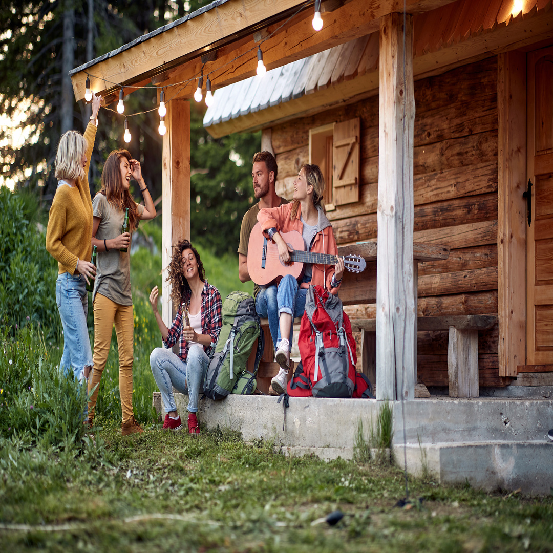 Six friends relax on a rustic cabin porch, playing guitar, laughing, and enjoying drinks in a forested outdoor setting.
