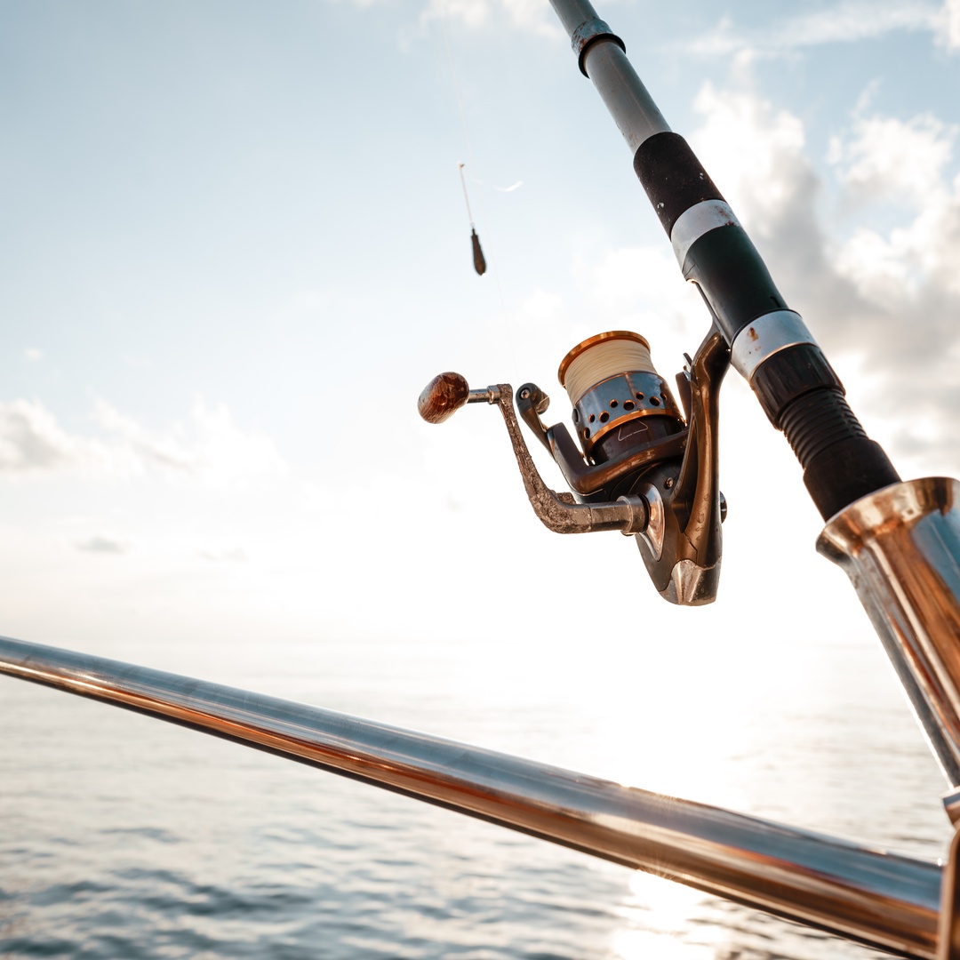 A fishing rod with a metallic reel is secured to a boat railing, set against a bright sky and calm ocean in the background.