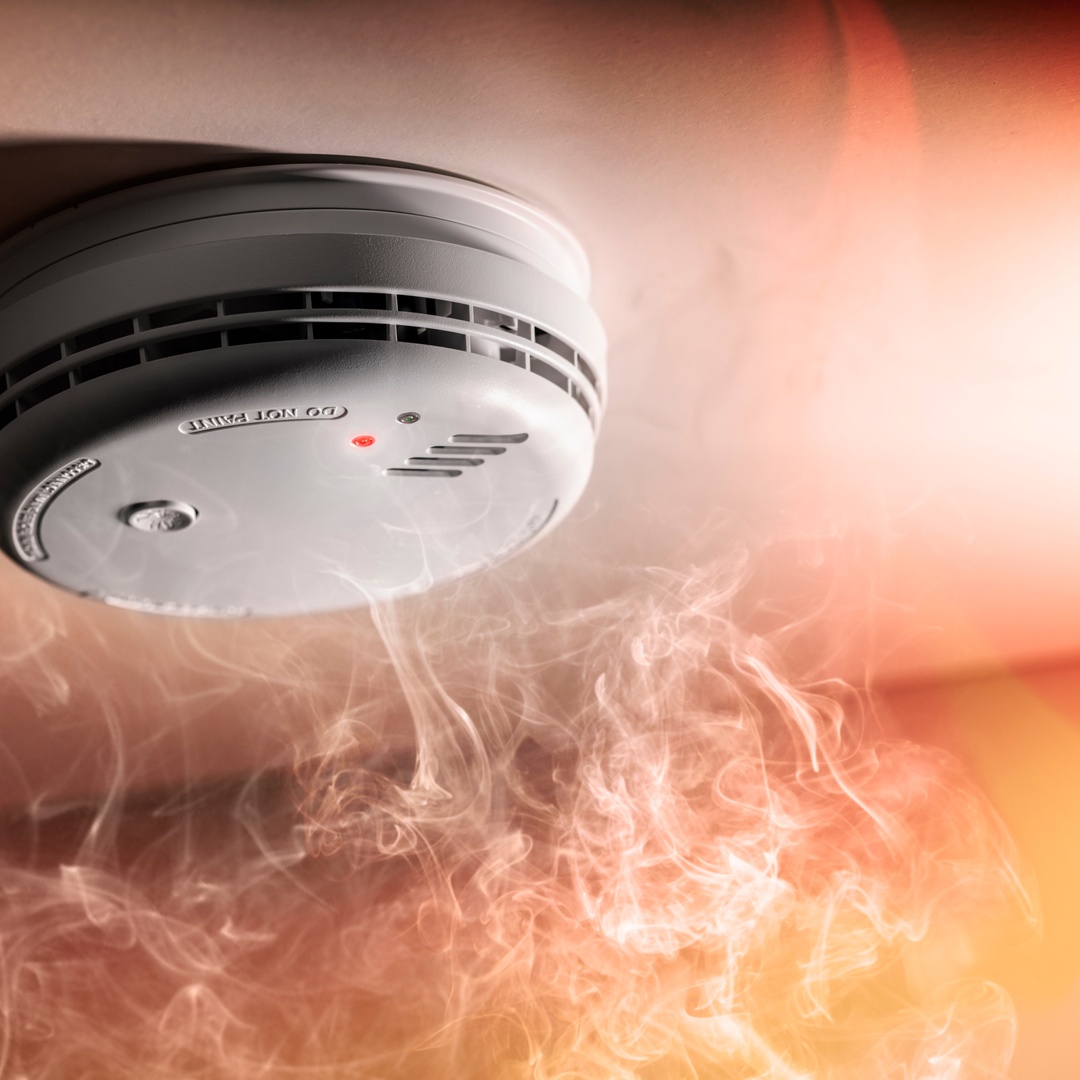 A smoke detector on a ceiling with a red light, surrounded by swirling smoke and an orange glow from a fire source.