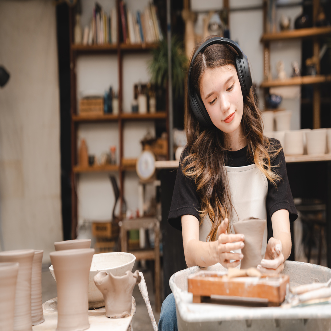 A woman working on a pottery wheel while wearing headphones. There is a wobbly piece of clay on her wheel.
