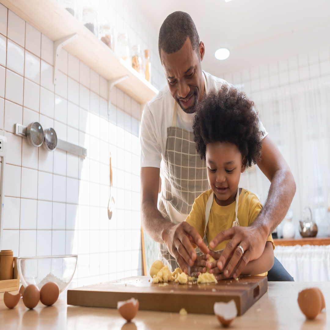 A father shows his son how to make cookies in the kitchen. Both are wearing aprons, and eggs sit on the counter.