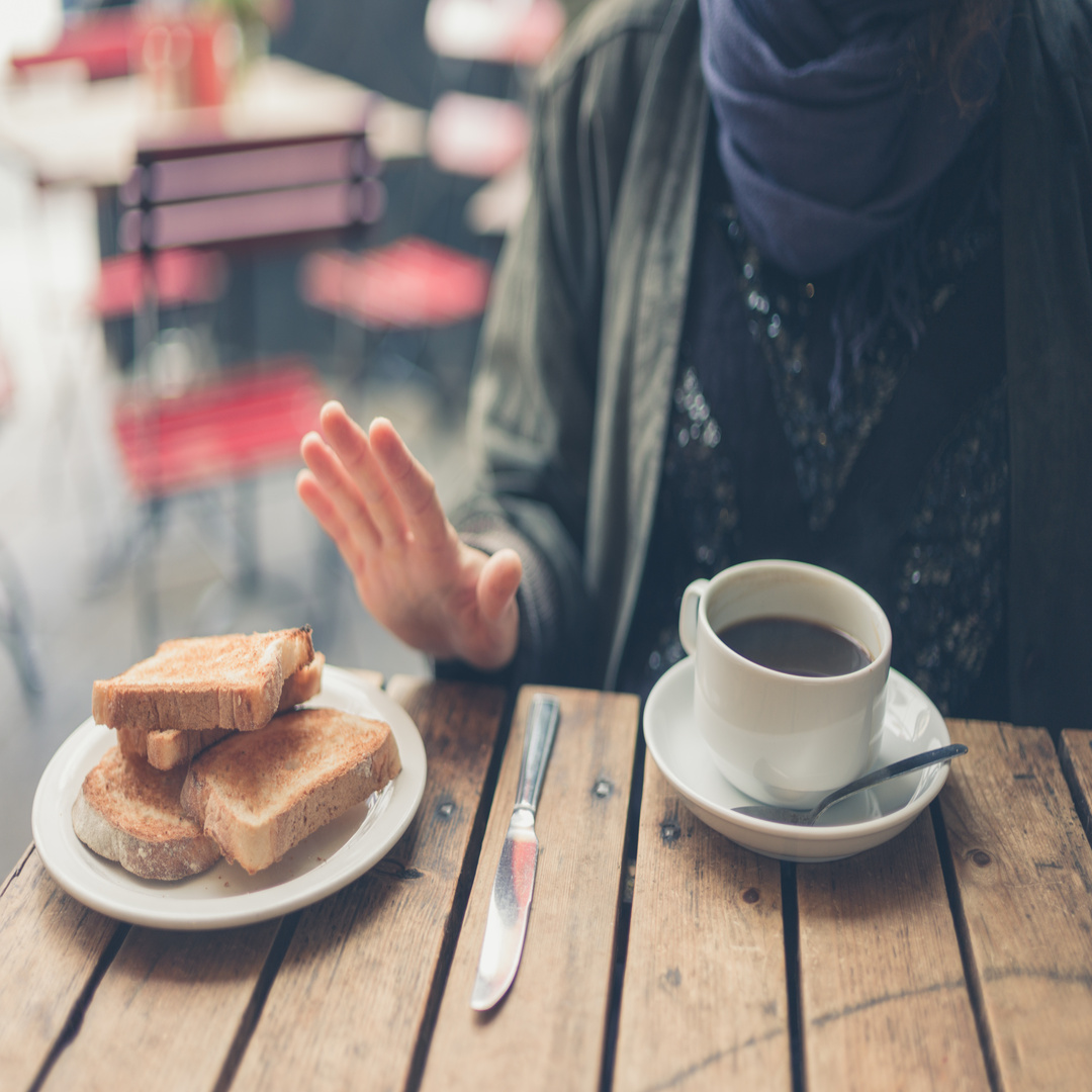 A close-up of a coffee cup and a plate of toast on a wooden table. Someone holds their hand up to push the toast away.