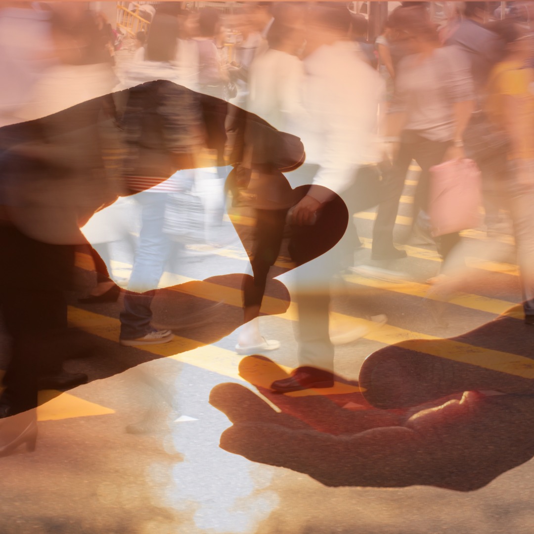 A close-up of a heart cutout passing between hands against a warm background, with a crowd shown in double exposure.
