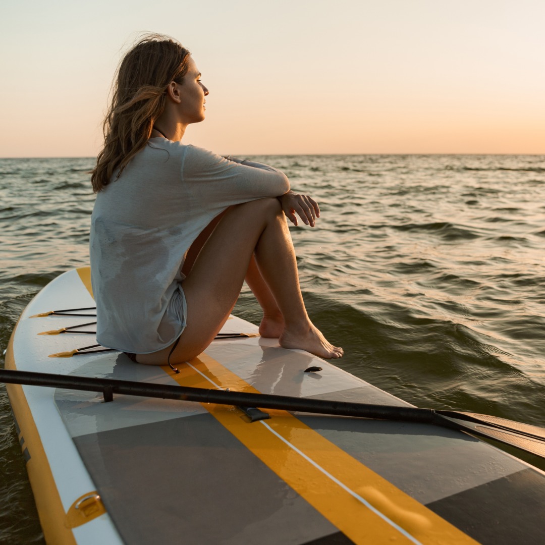 A twenty-something year old sits relaxed on her paddle board as she watches the sun set over the water.